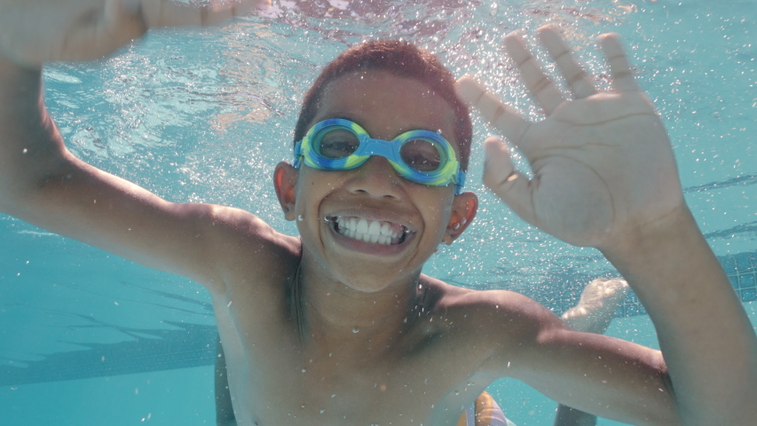 Funny little boy swimming underwater in pool smiling waving hand enjoying swim in crystal clear water wearing goggles enjoying summer 4k | Shutterstock HD Video #1034013797