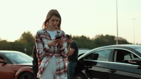 Confused girl at car crash using a smartphone calling for help standing with miserable expression outdoors. Road accident,. Police investigation.