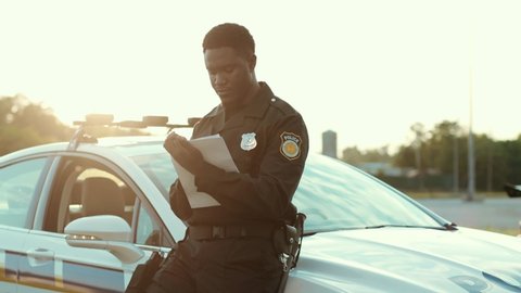 Portrait of attractive concentrated young black man police officer doing paperwork near the police car at sunlight. Police in the city.
