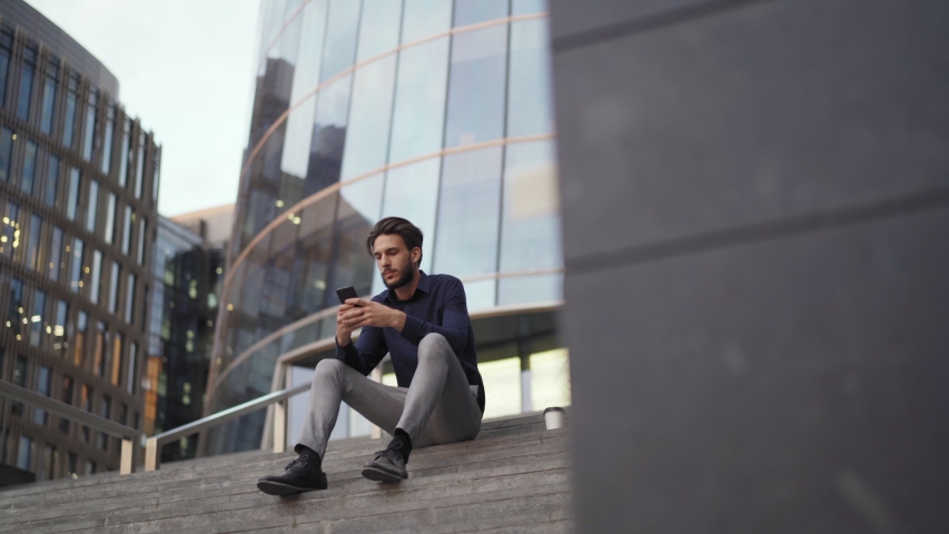 Camera approaching businessman sitting on steps of modern office building and text messaging on cell phone. Young man looking away dreamily Royalty-Free Stock Footage #1034017703