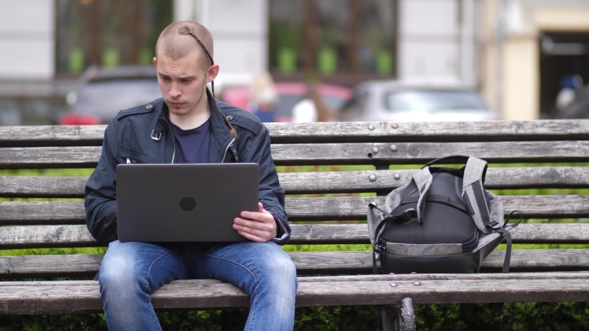 Focused male networking online on laptop pc sitting on bench in city park. Concentrated on work young student looking at screen of computer browsing Internet working remotely outdoor