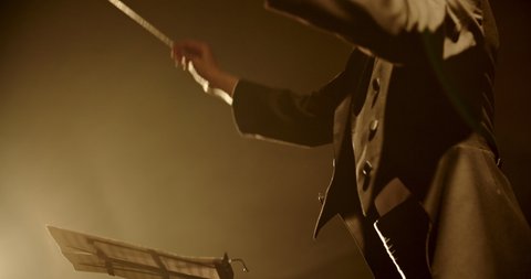 Unrecognizable male symphony orchestra conductor wearing black tux is directing musicians in orchestra pit by moving his hands and baton, studio shot on black background 4k footage