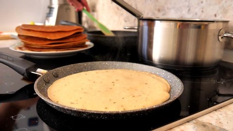 A female hand bakes traditional Russian tasty pancakes on an electric modern stove, overturns pancakes with a green silicone spatula. Cooking Russian pancakes. Slow motion.