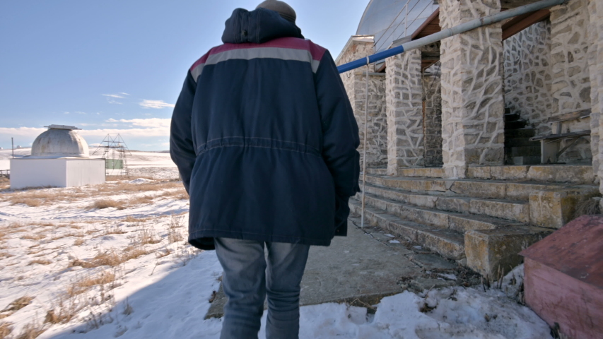 Following a male researcher in jeans and a warm uniform walking through the snow on a winter sunny day between the scientific buildings of the solar observatory. Scientific research station