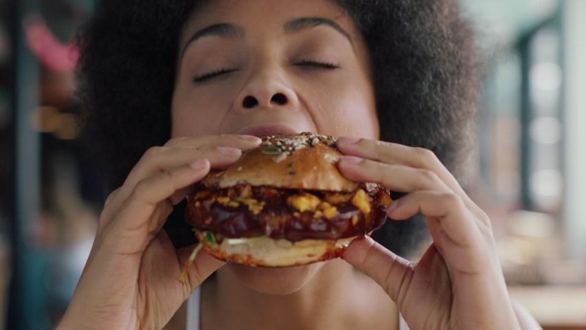 Beautiful woman with afro eating burger in restaurant enjoying delicious juicy hamburger mouth watering meal african american female having lunch 4k | Shutterstock HD Video #1034053604