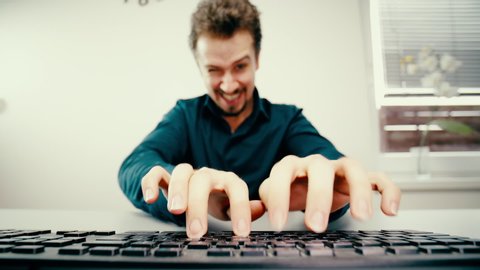 Man doing funny faces while typing on keyboard. Low angle view from behind the keyboard with male person fingers in focus while typing.