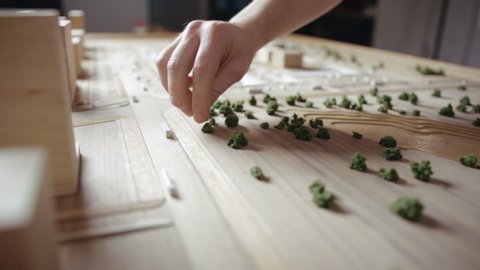 Person Decorates an Artificial Miniature Residential Quarter with Plants. Craftsman Arranges Decorative Shrubs with Fingers on a Wooden Layout. Caucasian Man Focused Addicting Hobby Close-up Shot