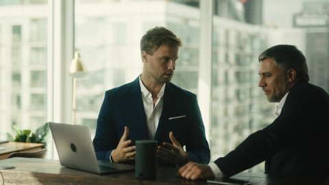Two businessmen discuss project details that are shown on laptop screen. Meeting in office with view on skyscrapers.