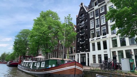 Footage of boat house on canal, historical and traditional buildings, trees in Amsterdam. It is a sunny summer day. Camera moves forward.