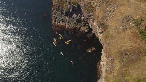 Cinematic aerial birds eye shot of kayaks in ocean by big cliffs. Sunny day, water shimmering. Turning shot.