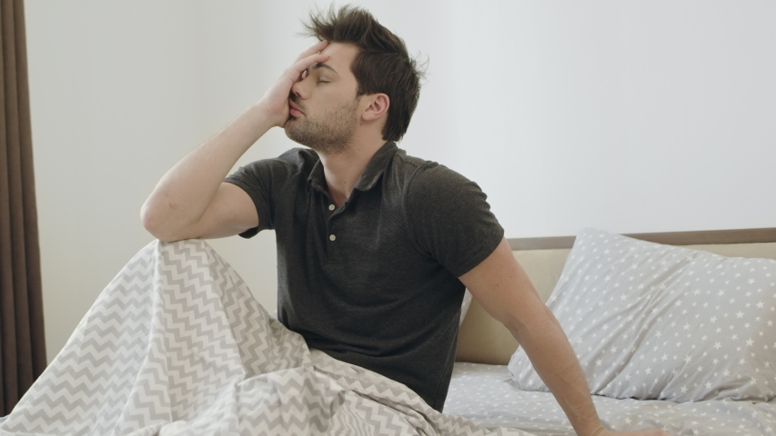 Drunk man trying to remember party night. Sleepy man waking up after party in bedroom in morning. Upset man feeling bad sitting in bed. Royalty-Free Stock Footage #1034089034
