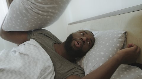 Upset man lying in bed in morning. African guy covering head with pillow at bedroom. Yound guy hating to wake up. Male person swearing in bed.