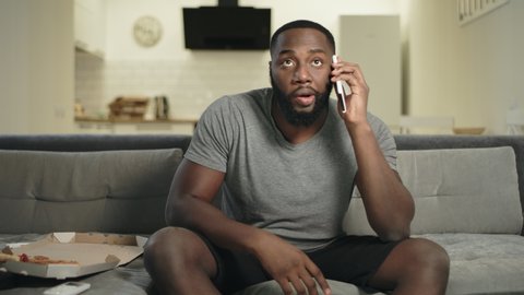 Black man eating pizza in front of tv at home. Focused man watching tv and talking mobile phone. Male fan commenting match results by cellphone.