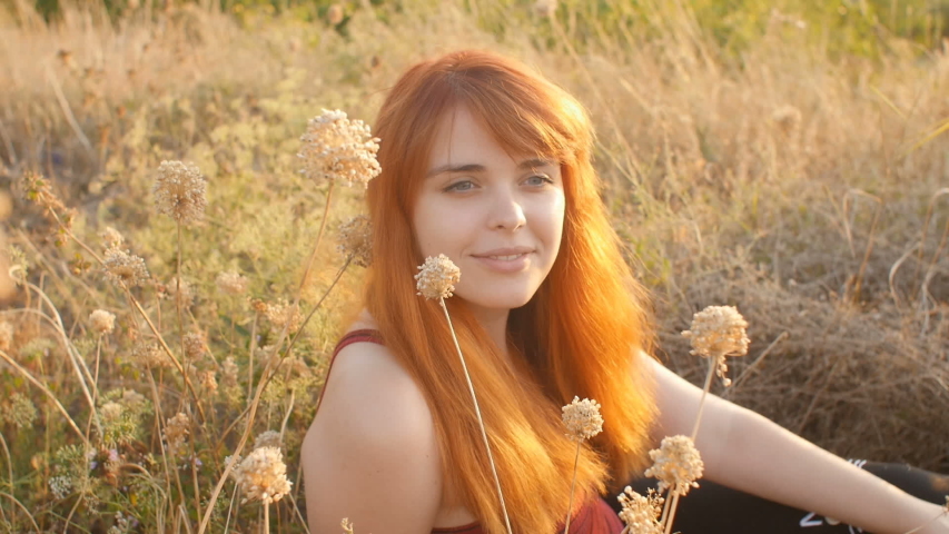 beautiful red-haired girl sits on the ground in a field among dried flowers and enjoys nature at sunset, young woman relaxing, concept of rest, healthcare, harmony, lifestyle