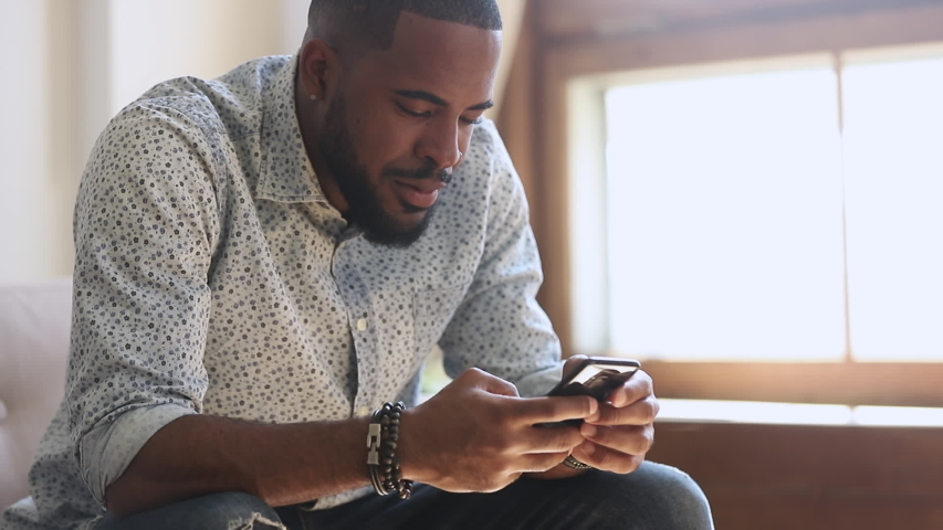 Young african american man holding smartphone device texting sms message sitting at home office, smiling black guy using apps playing mobile game chatting in social media surfing web on phone indoors Royalty-Free Stock Footage #1034099156