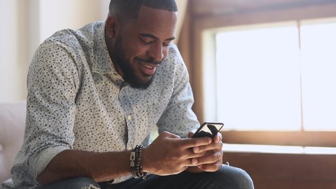 Young african american man holding smartphone device texting sms message sitting at home office, smiling black guy using apps playing mobile game chatting in social media surfing web on phone indoors
