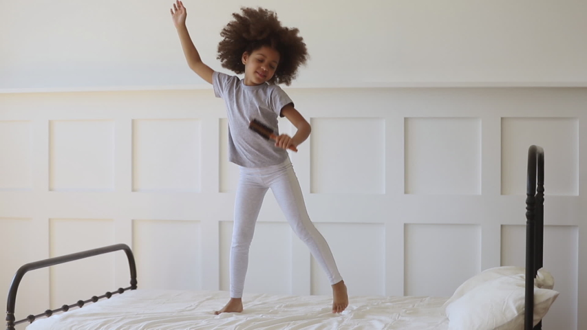 Funny adorable happy kid girl jump on bed sing in hairbrush microphone, cute little african american child having fun dancing to music playing in bedroom alone enjoy pretending singer in the morning Royalty-Free Stock Footage #1034099159