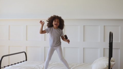 Funny adorable happy kid girl jump on bed sing in hairbrush microphone, cute little african american child having fun dancing to music playing in bedroom alone enjoy pretending singer in the morning