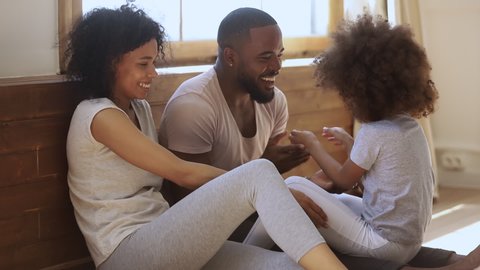Happy black family mom dad and little cute child daughter laughing tickling sit on bedroom floor, cheerful parents having fun with small funny african kid playing cuddling bonding together in morning