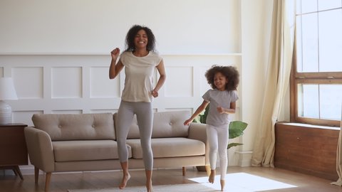 Active happy african mom and cute little kid daughter jumping dancing at home, carefree funny black family mother having fun with small child girl laughing do morning exercise together in living room