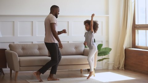 Happy funny carefree african family father and cute little kid daughter jumping having fun together in living room, dad dancing with small child girl enjoy music active morning playing at home