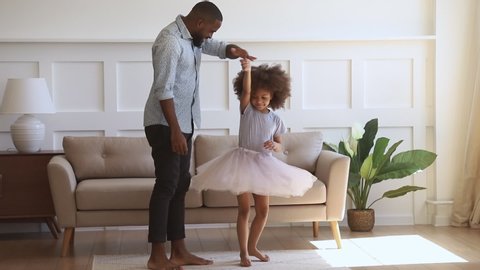 Happy family young african american dad holding hand of child daughter dancing with his small princess standing on knee kissing hand thanking cute little kid girl for dance playing in living room