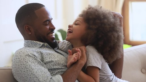 Happy african american family young father and cute small child daughter bonding embracing tickling sit on sofa, loving black dad playing with little kid girl hugging laughing cuddling at home