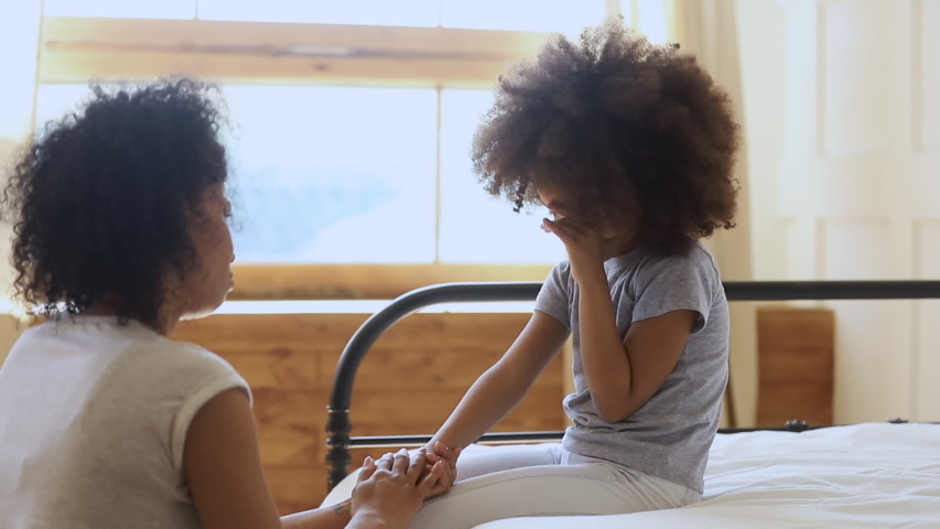 Worried loving african mother holding hand comforting upset little bullied kid daughter crying talking give support or apologizing, mom foster parent helping small child with problem sit on bed | Shutterstock HD Video #1034099264