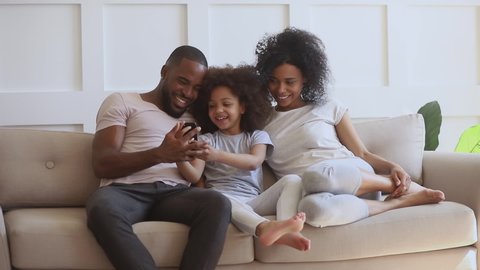 Happy african family with small cute kid daughter sit on sofa having fun with smart phone, little child girl holding cellphone using funny mobile app laughing with black parents take selfie at home