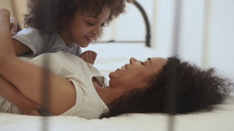Cute funny little african american child daughter tickling happy mother playing together lying on bed, cheerful mixed race family small kid girl and black mom embrace cuddle laugh relax in bedroom