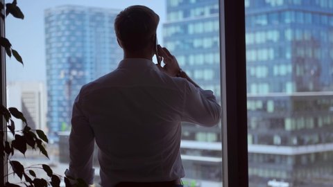 A stylish businessman walks to the window of his office, answers the phone and calls and discusses the business plan against the background of high-rise buildings and a business center.