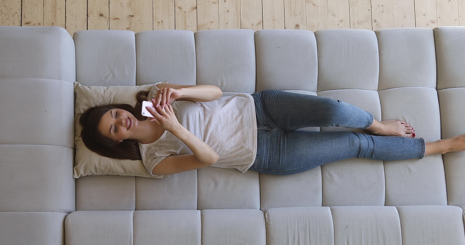 Happy relaxed girl holding smart phone using mobile apps watching funny video laughing lying on couch, smiling lazy young woman having fun chatting in social media resting on sofa at home, top view Royalty-Free Stock Footage #1034100872