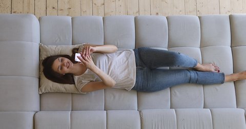 Happy relaxed girl holding smart phone using mobile apps watching funny video laughing lying on couch, smiling lazy young woman having fun chatting in social media resting on sofa at home, top view