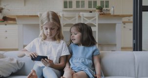Two funny cute girls sisters learning using smartphone taking selfie at home, little kids siblings holding phone enjoy mobile social media app play game sit on sofa, children and technology concept