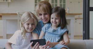 Happy young mother and cute little daughters taking selfie watching cartoons on smartphone, smiling mom and children girls using funny mobile app on cellphone look at screen laughing bonding at home