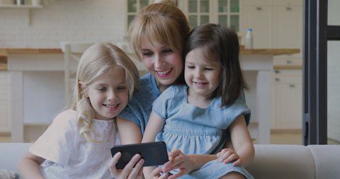 Happy young mother and cute little daughters taking selfie watching cartoons on smartphone, smiling mom and children girls using funny mobile app on cellphone look at screen laughing bonding at home