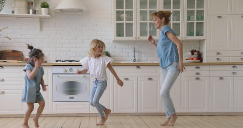 Happy family active young mom and two cute small children dancing jumping together in kitchen, carefree funny little daughters copy imitate mother baby sitter having fun do exercises laughing at home