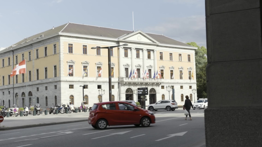 A building with many flags on a busy street with moving cars, embassy in Switzerland. Action. A street with driving vehicles near the beige municipal building with european flags.