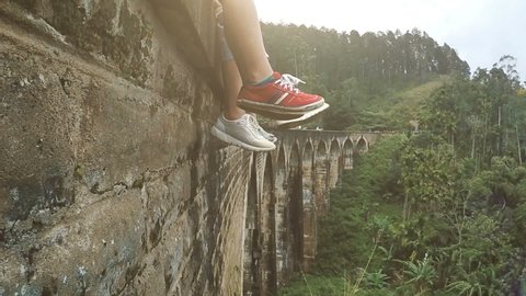 4K footage of two people's feet in sport shoes dangling over valley. People sitting on stone viaduct in tropical forest.