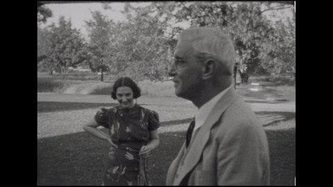 1930s: Elderly man and younger woman on front lawn, man walks off, woman smiles and points. Woman bends over grass. Baby girl in frilly dress runs down stone walkway. Woman pushes older girl in swing.