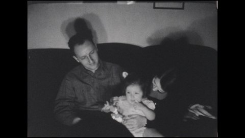 1930s: Man and woman sit on couch with baby girl, man shows her a toy duck, baby grabs it, puts it in her mouth, man looks at camera. Woman stands up baby on her leg, smiles at camera.