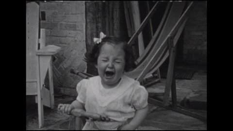 1930s: Baby girl screams, holds tricycle handle. She stands by self on lawn, clutching her hands. She plays with cardigan. She stands on lawn in romper. Baby squats, plays with sprinkler propeller.