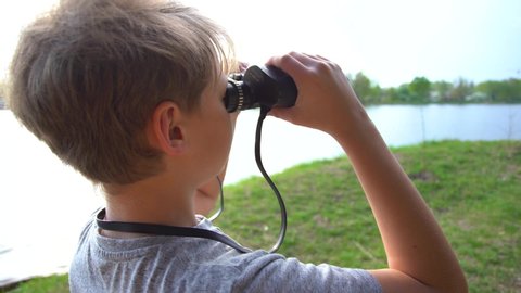 Closeup portrait of cute curious kid exploring nature using old vintage binoculars. Boy isolated on blurry river water and green meadow background. Real time 4k video footage.