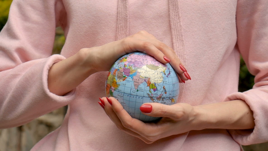 Young female teenager in pink casual clothes with red manicure holding a little globe with geographical names in Ukrainian cyrillic letters on it in her hands. Enviromental responsibility concept.