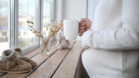 Pregnant Woman Holding Cup Of Tea And Touching Her Belly At Home.