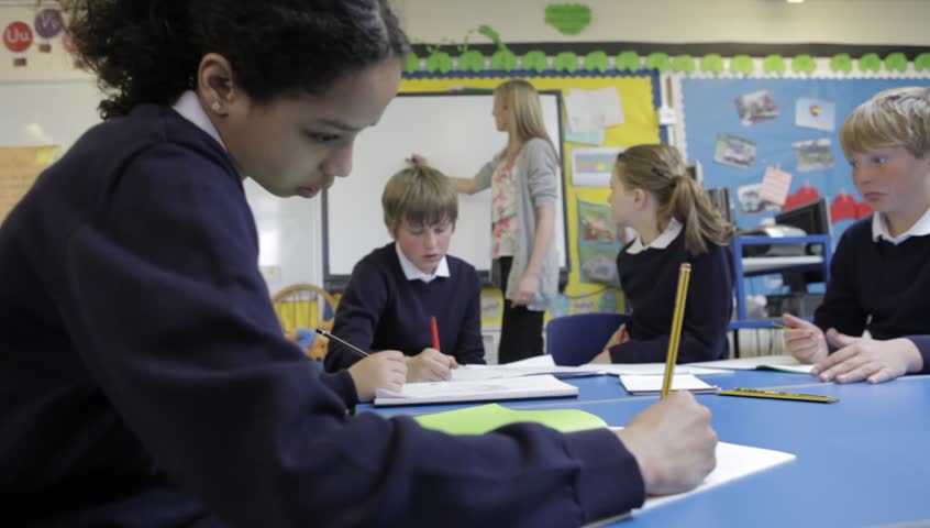 Children working at desk writing in books with teacher helping in background.Shot on Canon 5d Mk2 with a frame rate of 30fps.