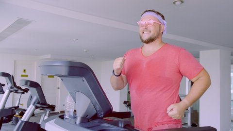 Funny fat male in pink glasses and in a pink t-shirt is engaged on a treadmill in the gym depicting a girl. 4k.