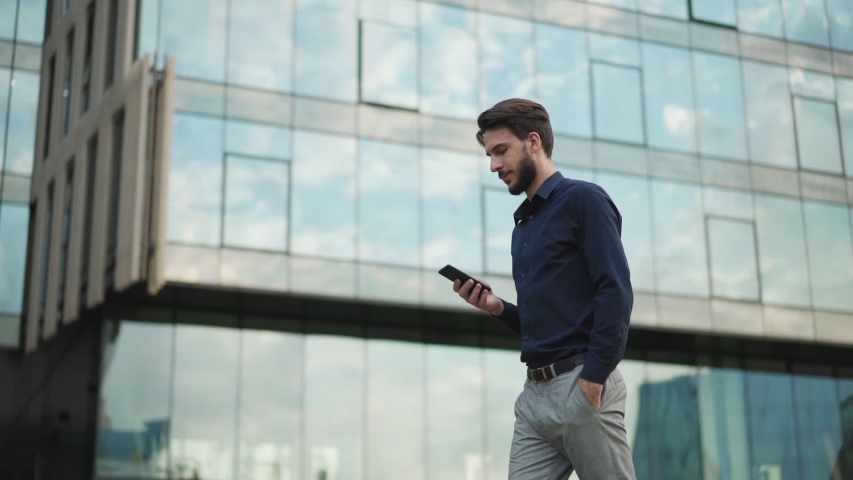 Tracking zoom in shot of handsome young businessman text messaging on cell phone while walking down city street in slow motion with hand in pocket. Man looking forward and away thoughtfully Royalty-Free Stock Footage #1034148035