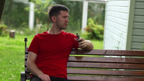 Vape man. Young caucasian white guy in red shirt sitting on a bench, smoking and letting off steam from an electronic cigarette in the backyard in the summer. Bad habit. Vaping activity. Lifestyle.