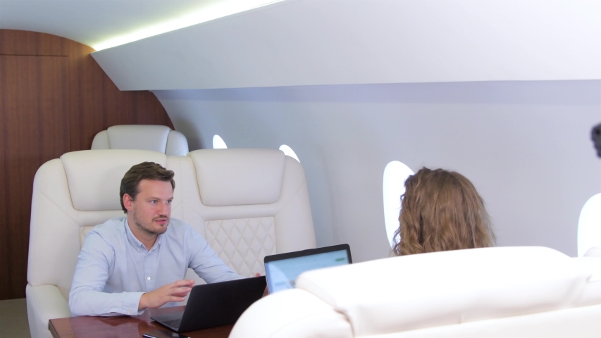 Work on laptop on board of private jet. Biracial flight attendant offering and pouring glass of wine for caucasian businessman and businesswoman travel inside of business airplane cabin. Royalty-Free Stock Footage #1034149367